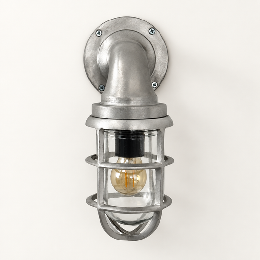 Industrial-style wall sconce with a brushed silver metal mounting plate, elbow pipe, cage guard, and visible Edison bulb on a plain off-white wall.