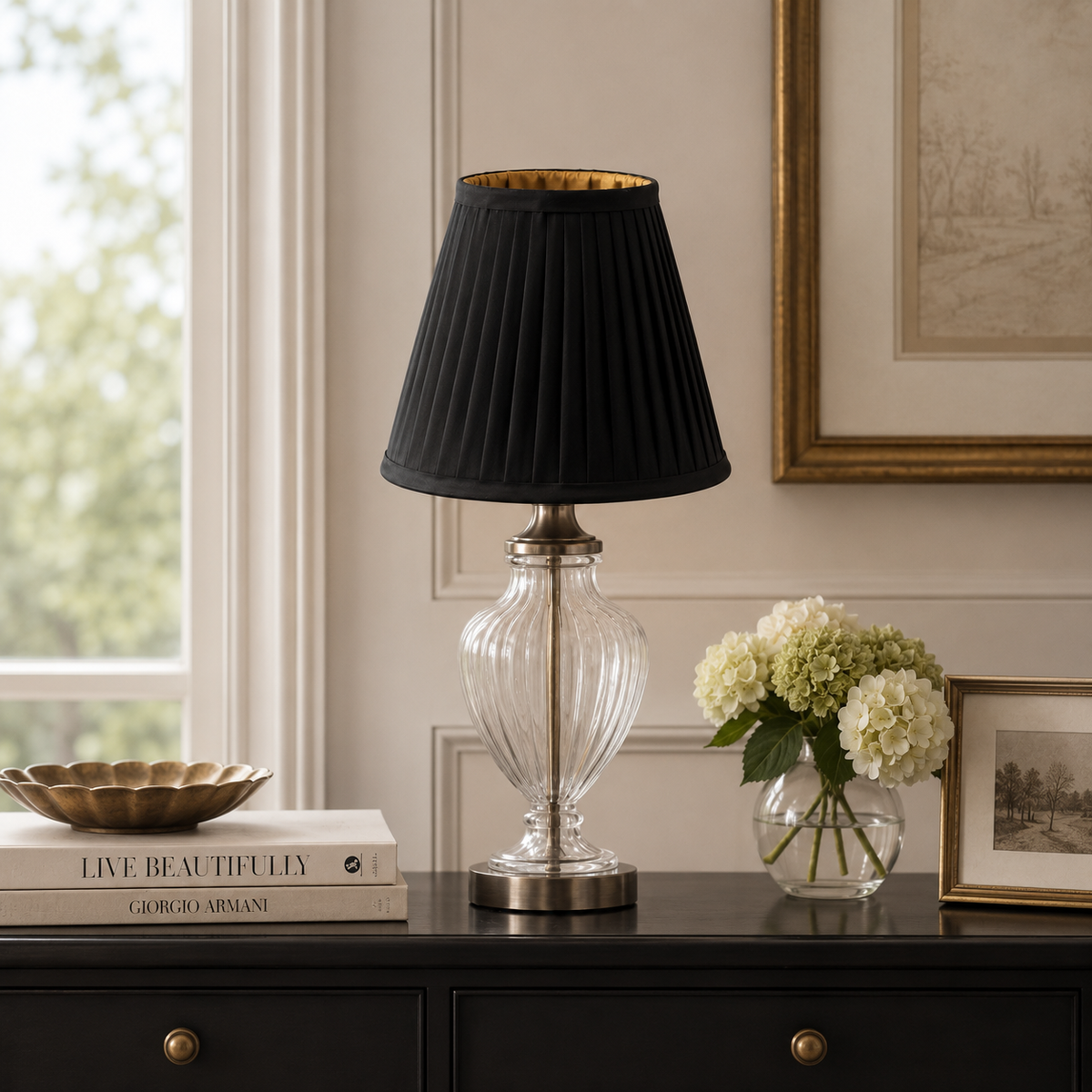 A table lamp with a black pleated fabric shade, a clear ribbed glass base, and a brushed silver metal foot ring, styled on a dark wooden console table in a decorated room.