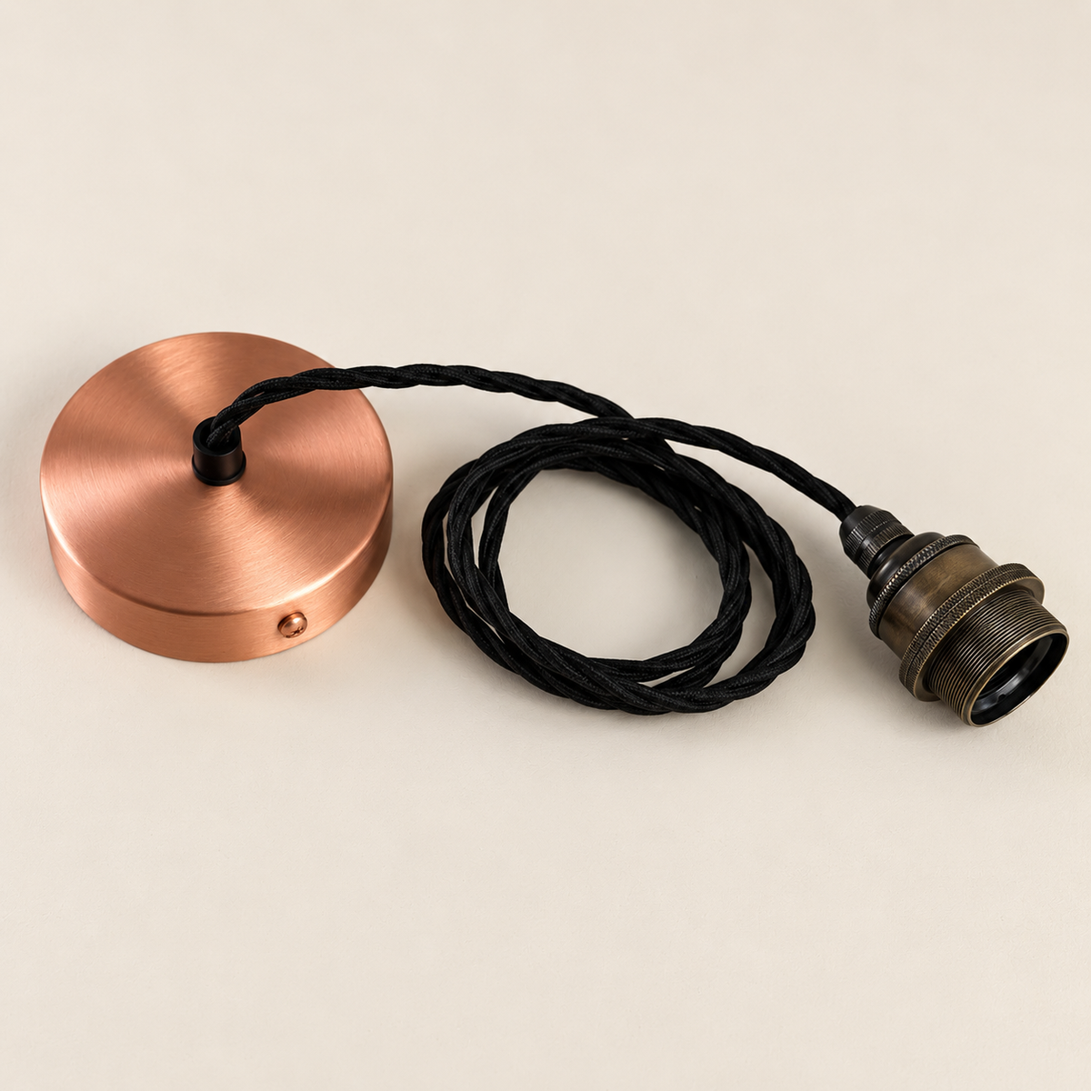 A brushed copper circular ceiling mount with a black braided cord and an antique bronze socket, photographed on a warm off-white background.