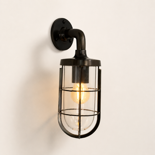 Industrial-style wall sconce with antique brass metal wall mount, curved arm, and clear glass cylindrical cage housing a glowing Edison bulb against a plain off-white wall.