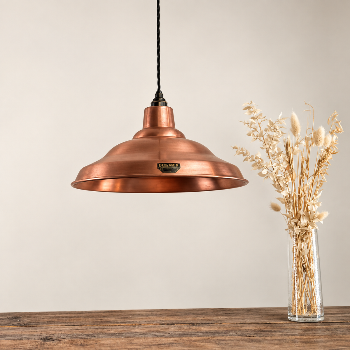 A copper-colored metal pendant light with a black cord hangs over a rustic wooden table next to a clear glass vase filled with dried grasses, against a neutral beige background.