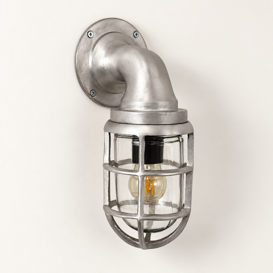 Industrial-style wall sconce with a brushed silver metal pipe mount, protective cage, and visible Edison bulb, shown against a plain off-white wall.