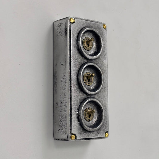 A rectangular metal light switch with a distressed silver finish, featuring three circular recessed sections on top with small brass-colored knobs or centers, photographed on a plain light background.