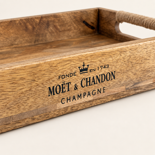 A close-up of a wooden serving tray with a rope handle, featuring a black printed Moët & Chandon Champagne logo on the front, set against a clean off-white background.