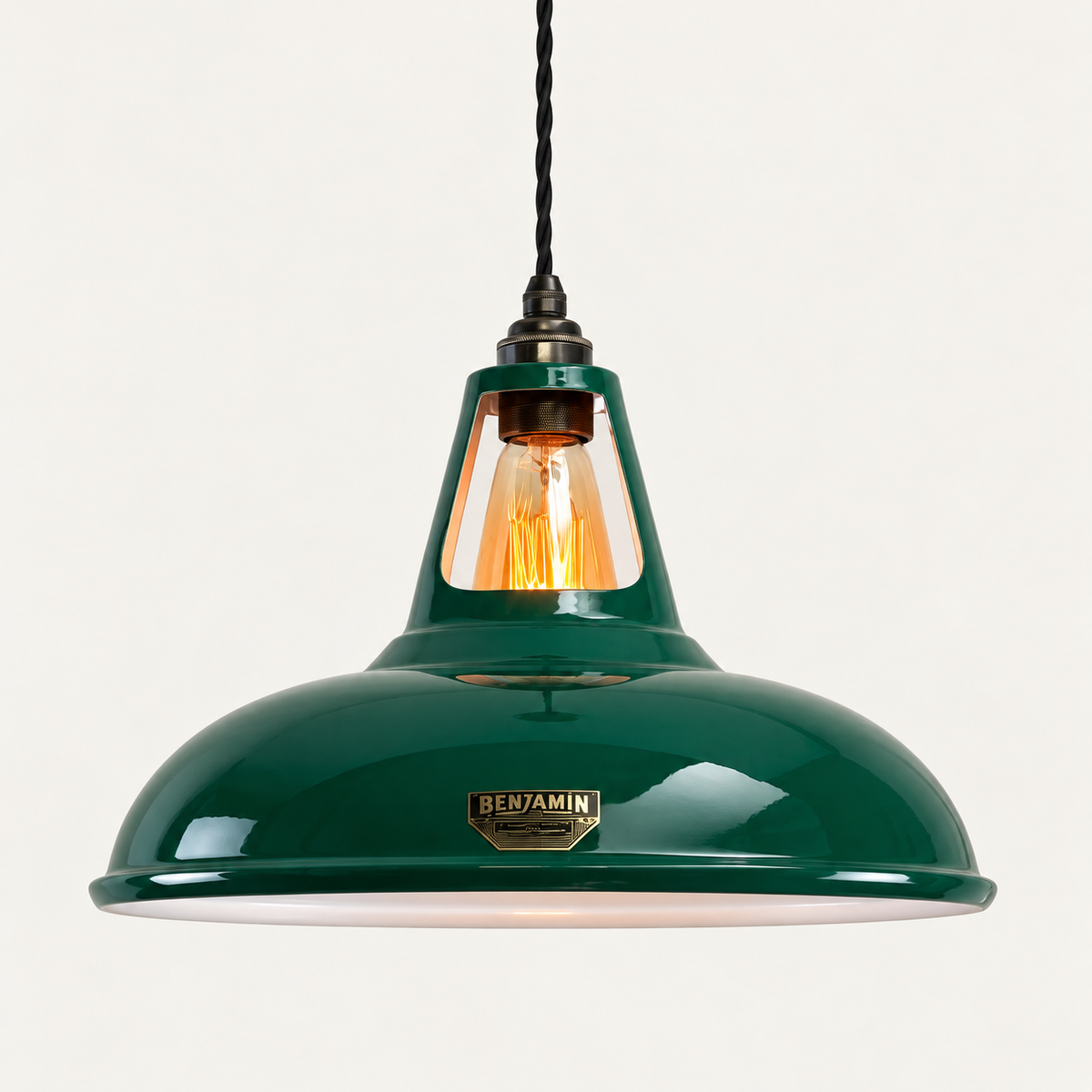 A green pendant light with a glossy enamel finish, featuring a brass 'BENJAMIN' brand plate on the front of the shade. The light is shown illuminated with a warm glow from the visible filament bulb, suspended by a black cord against a clean, off-white background.