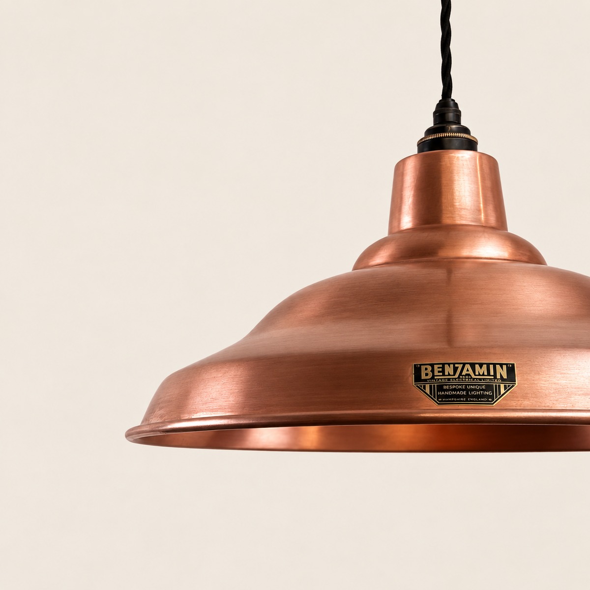 A close-up of a copper pendant light shade by Benjamin, showing the brushed metal finish and a black hanging cord against a warm off-white background.