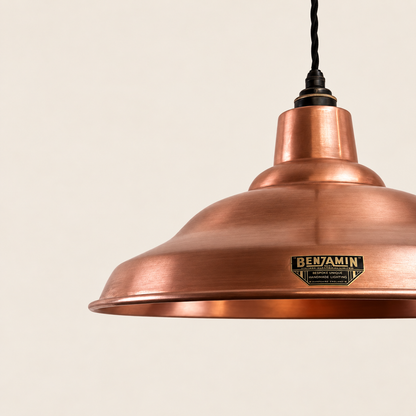 A close-up of a copper pendant light shade by Benjamin, showing the brushed metal finish and a black hanging cord against a warm off-white background.