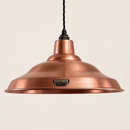 A copper pendant light with a wide, shallow dome shade, a black hanging cord, and a small metallic Benamin brand plaque on the front, set against a solid off-white background.