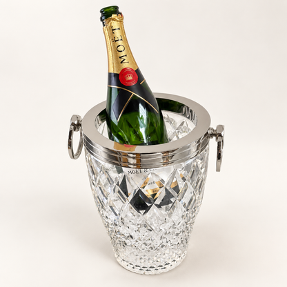 A Moët sparkling wine bottle in a green glass bottle with a gold foil capsule, placed inside a clear cut-crystal champagne bucket with silver metal handles, on a clean white background.
