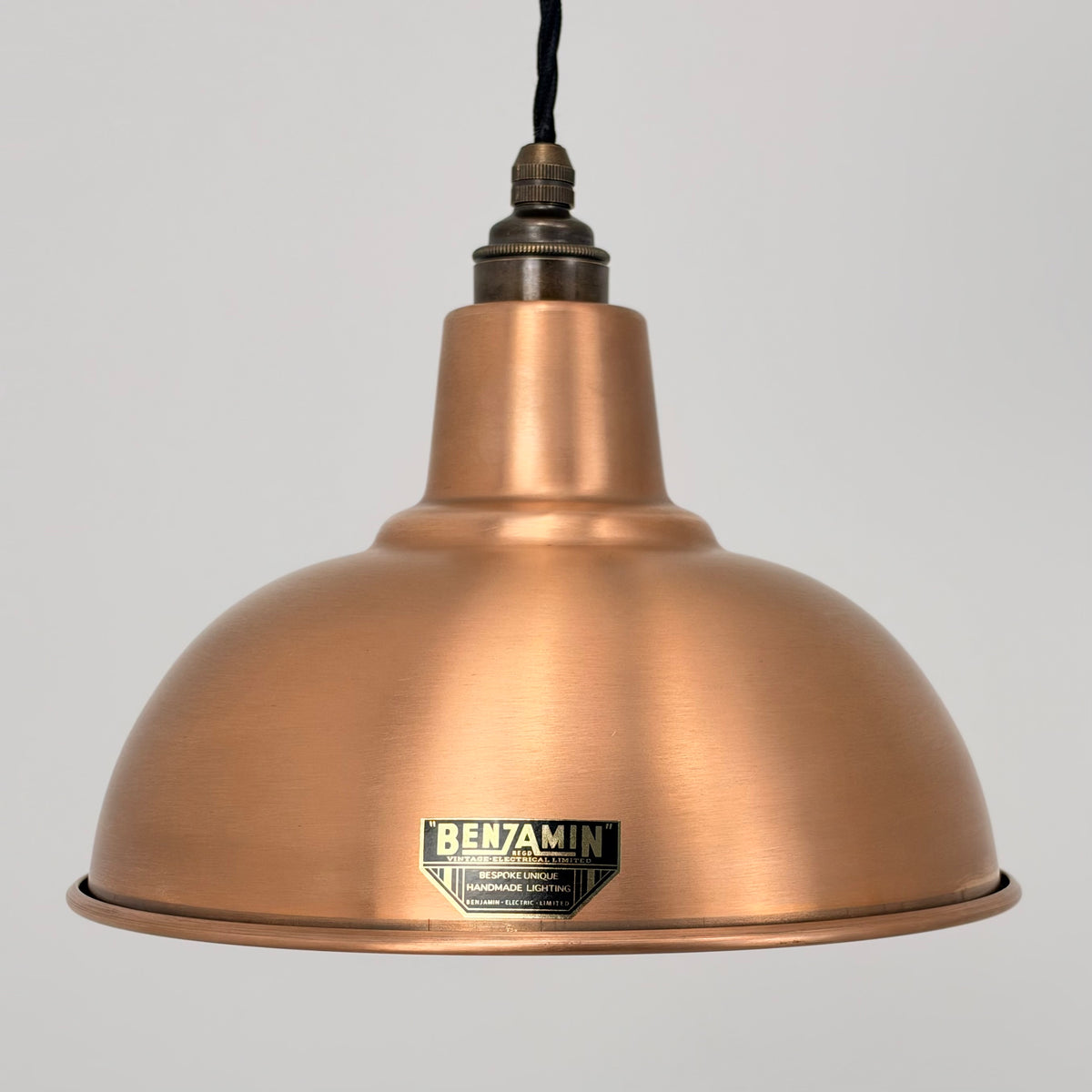 A copper-colored metal dome pendant light shade with a black hanging cord and a visible Benjamin brand badge on the front, set against a plain light gray background.