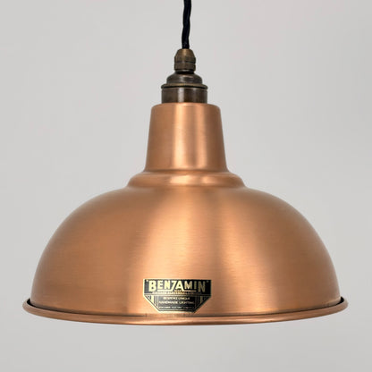 A copper-colored metal dome pendant light shade with a black hanging cord and a visible Benjamin brand badge on the front, set against a plain light gray background.