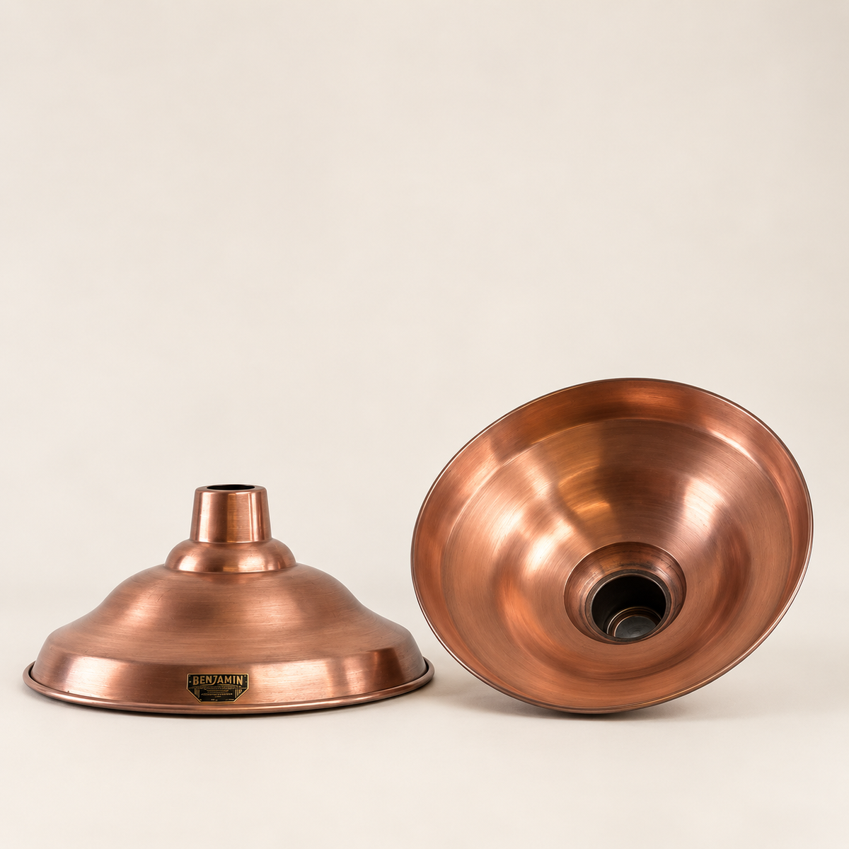 Two copper-colored metal lamp shades shown on a plain light beige studio background, one upright and one tipped on its side to show the interior.