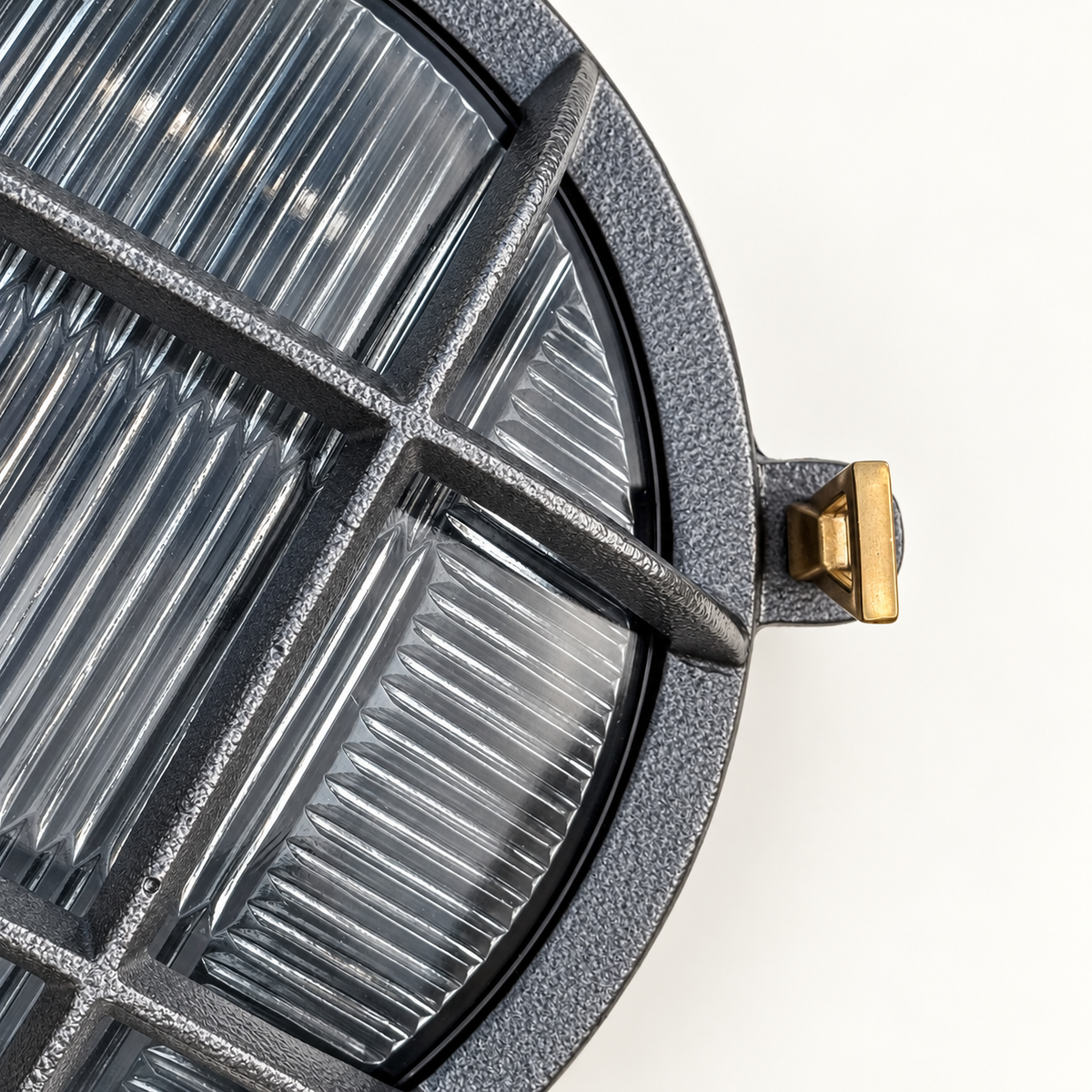 Close-up of a round outdoor light fixture with a textured dark grey frame, ribbed clear lens, and a small brass mounting tab on a white background.