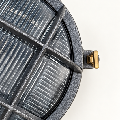 Close-up of a round outdoor light fixture with a textured dark grey frame, ribbed clear lens, and a small brass mounting tab on a white background.