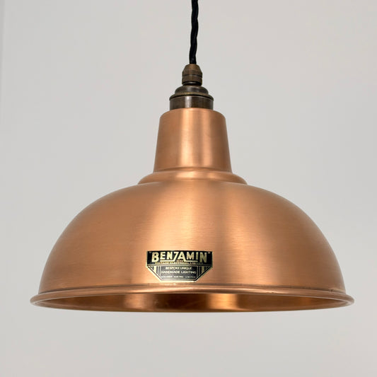 A copper dome pendant light with a black hanging cord and a visible BENJAMIN brand label on the front, set against a plain light gray background.