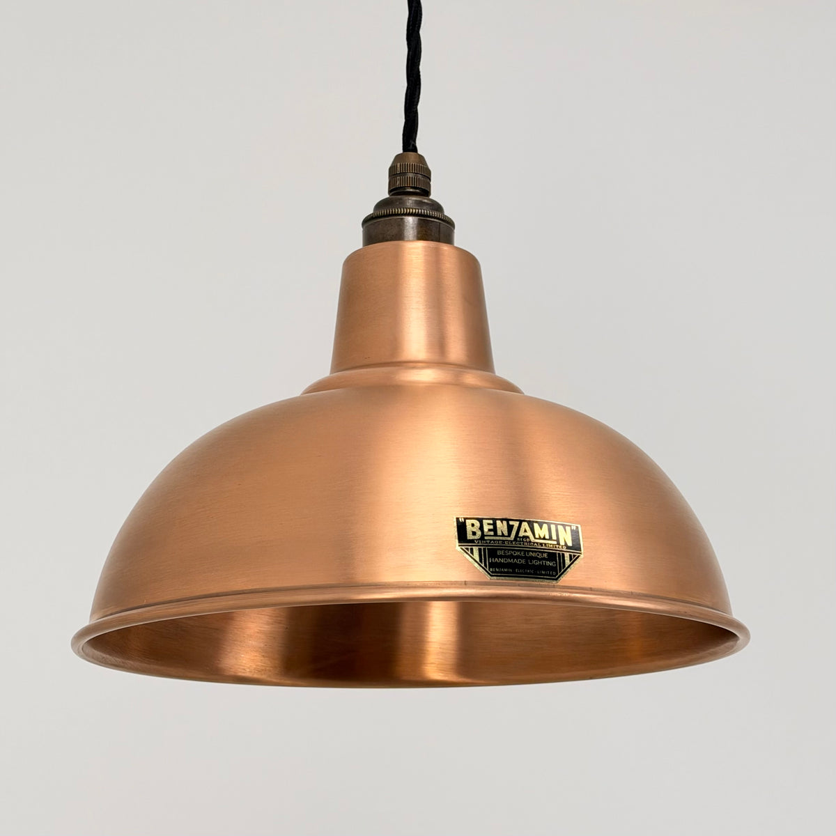 A copper-colored metal pendant lamp shade with a black hanging cord and a visible BENJAMIN brand badge on the front, set against a plain light gray background.