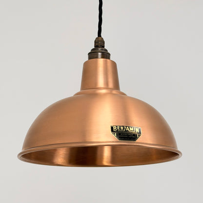 A copper-colored metal pendant lamp shade with a black hanging cord and a visible BENJAMIN brand badge on the front, set against a plain light gray background.