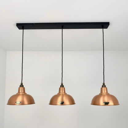 Three copper-colored pendant light shades hanging from a black ceiling bar with black cords, shown against a plain white wall and ceiling.