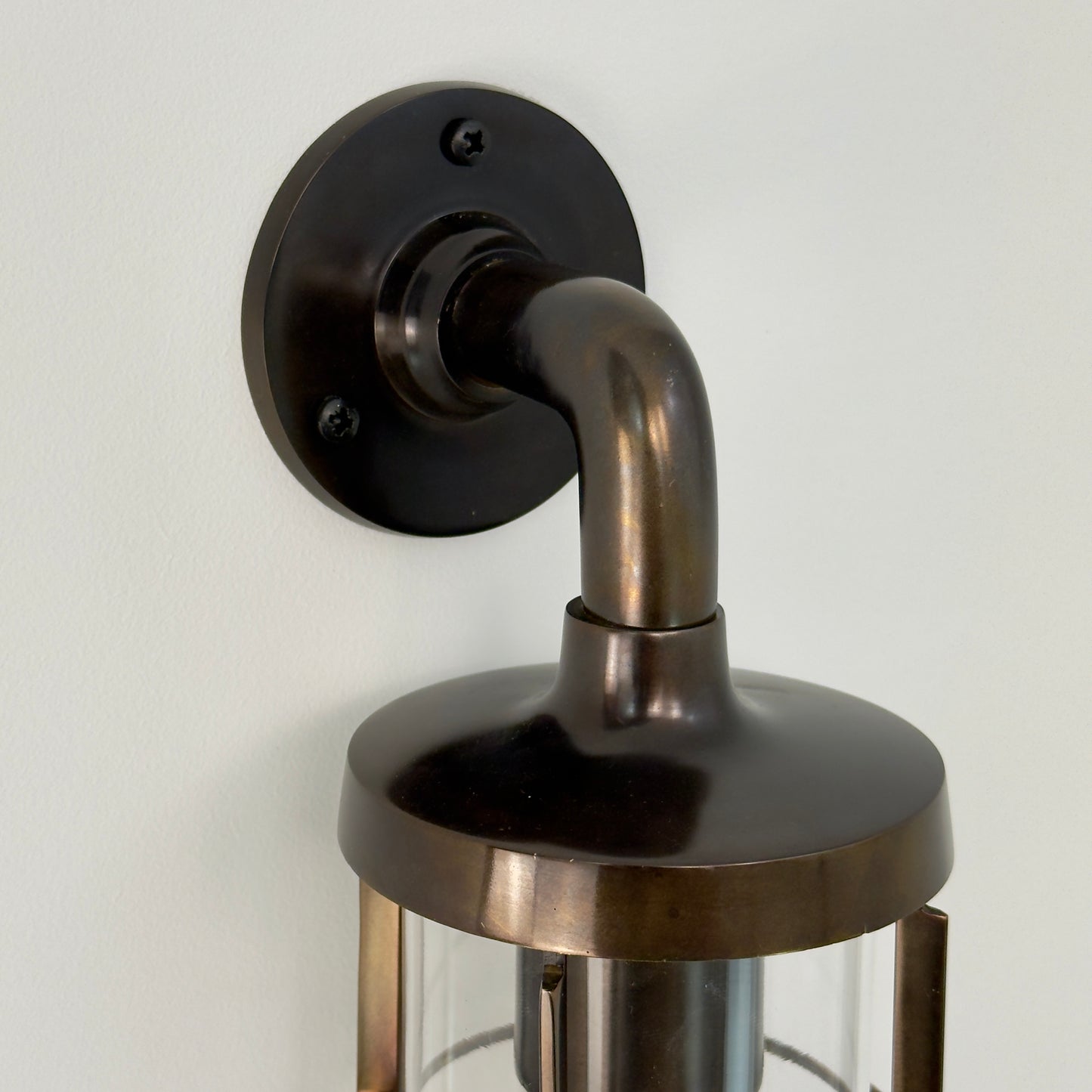 A close-up of a wall-mounted lantern light fixture with an antique genuine solid brass body, curved neck, round wall plate, and glass shade, shown against a plain off-white wall.