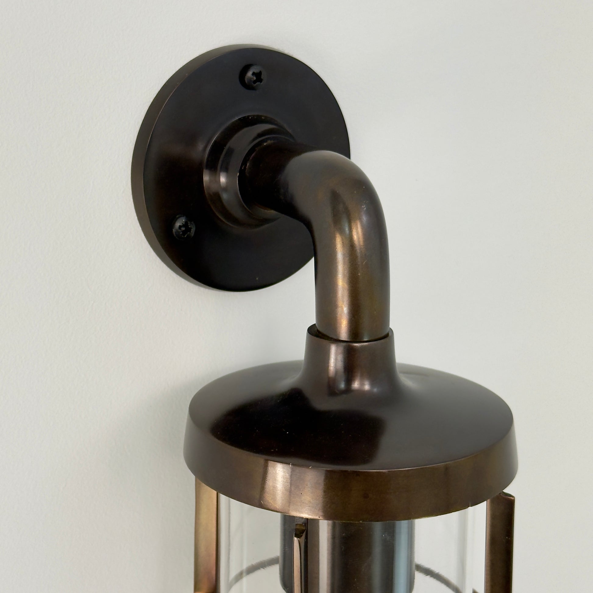A close-up of a wall-mounted lantern light fixture with an antique genuine solid brass body, curved neck, round wall plate, and glass shade, shown against a plain off-white wall.
