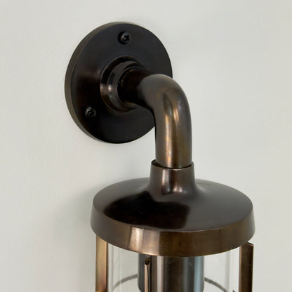 A close-up of a wall-mounted lantern light fixture with an antique genuine solid brass body, curved neck, round wall plate, and glass shade, shown against a plain off-white wall.